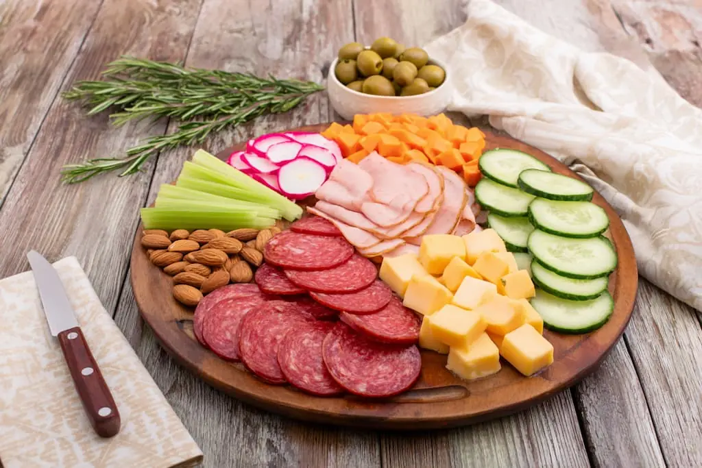 Wooden charcuterie board featuring keto-friendly deli meats including salami and ham, aged cheddar cheese cubes, green olives, almonds, cucumber slices, and fresh vegetables on rustic wood background