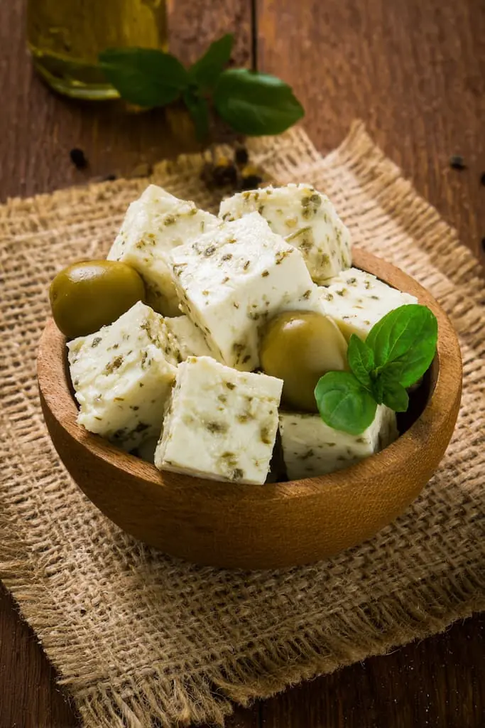 Herb-crusted white cheese cubes with green olives in wooden bowl for keto Mediterranean snacking