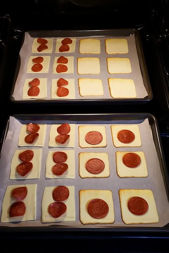 Two baking sheets with cheese squares topped with pepperoni arranged on parchment paper ready for oven