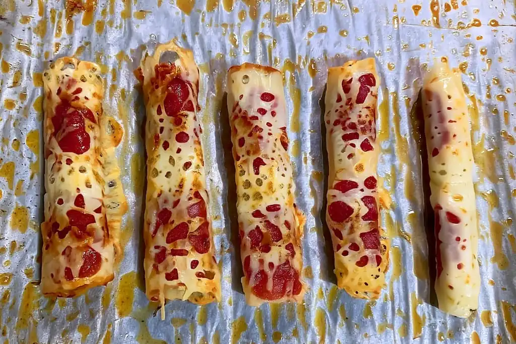 Five rolled keto pizza roll-ups with golden cheese and pepperoni pieces on parchment paper