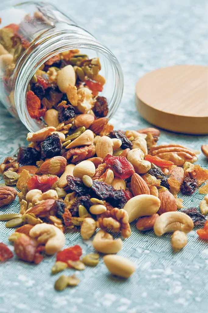 Mixed nuts and dried fruit trail mix spilling from glass jar onto blue kitchen towel