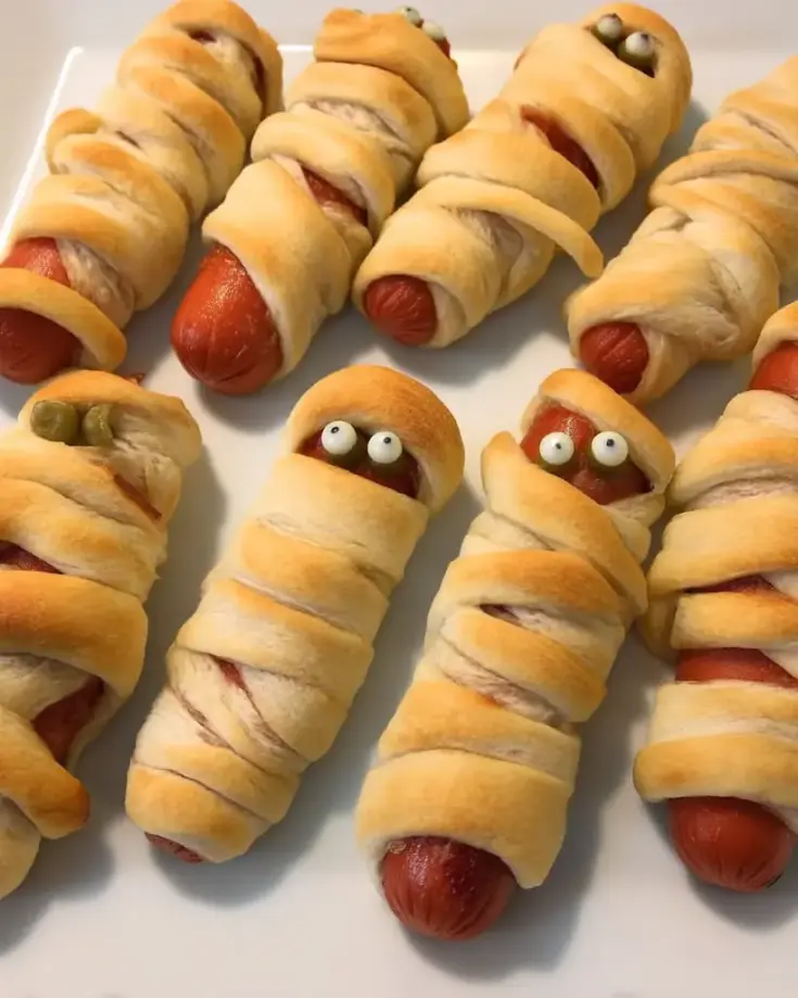 Golden brown mummy hot dogs wrapped in crescent roll dough with candy eyes on white plate