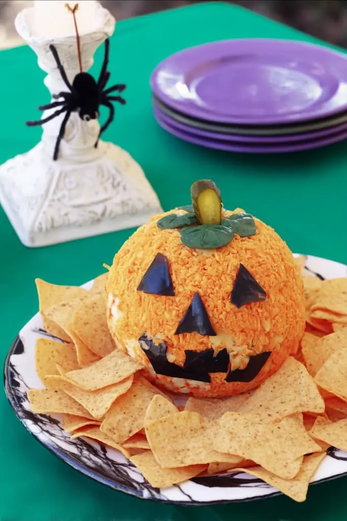 Jack-o'-lantern cheese ball with orange nacho chip coating, black olive face, and pickle stem surrounded by tortilla chips on Halloween table