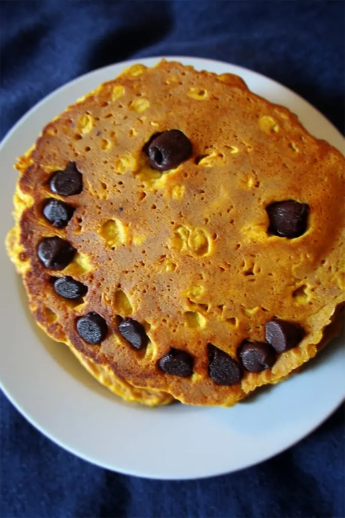 Overhead view of golden pumpkin pancake stack with chocolate chip jack-o-lantern smile face on white plate with dark blue background