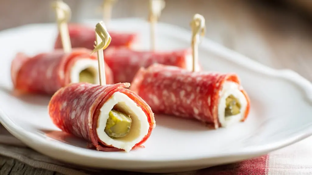 Keto salami roll-ups with cream cheese and pickles secured with toothpicks on white plate