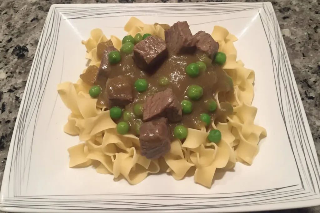 Close-up of cubed beef and bright green peas in a glossy sauce atop tender egg noodles, illustrating a sticky beef noodles recipe