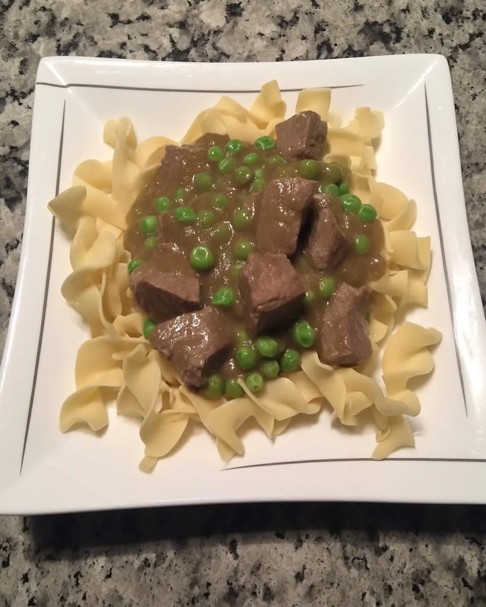 Cubed beef and bright peas coated in rich gravy over egg noodles on a white square plate—classic sticky beef noodles dish
