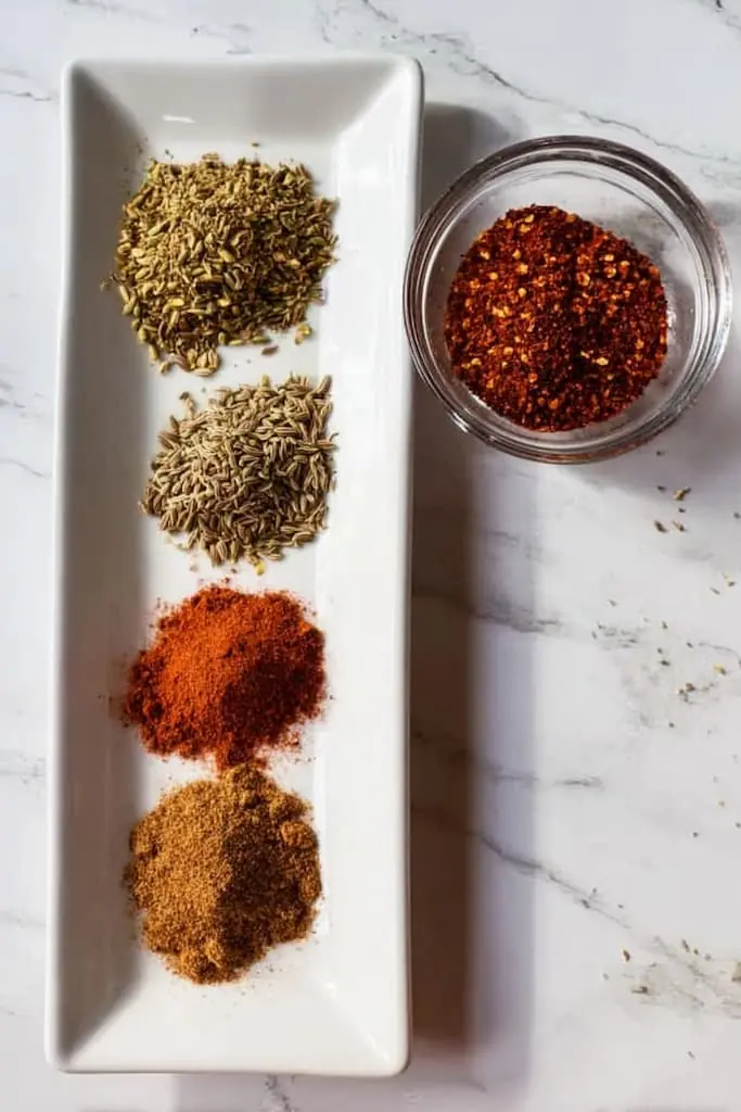Ground cumin, fennel seeds, oregano, and chili powder arranged on white plate with red pepper flakes in glass bowl