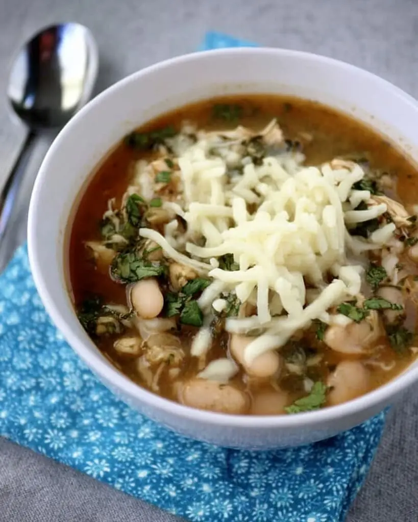 Bowl of white chicken chili topped with shredded cheese and fresh cilantro, showing tender chicken pieces and white beans in a clear broth
