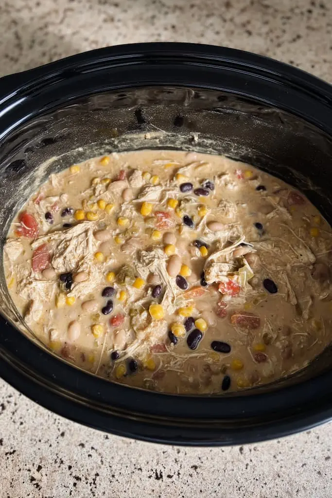 Creamy white chicken chili cooking in a black slow cooker with visible shredded chicken, white beans, black beans, corn, and diced tomatoes